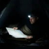 un homme lit une carte dans une tente de camping, éclaré par une lampe frontale qui éclaire en blanc